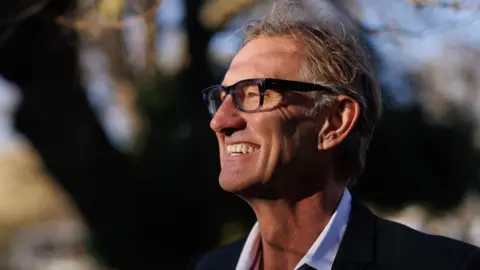 Tony Adams. A middle-aged man with thick black glasses and messy grey hair. He is smiling, looking off into the distance on the left and wearing a black suit jacket with white shirt