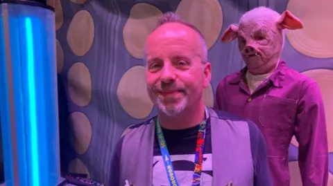 Sci-fi Scarborough's founder Steve Dickinson - a man with grey hair and a beard standing in front of a blue light and an extra-terrestrial model, which is wearing a pigs face mask covering their entire head and dark-coloured overalls.