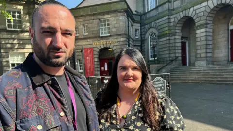 Ricky is nearer the camera. He has short hair and a black beard. He has a pattered coat on and is looking at the camera. Fiona is looking at the camera and has long black hair. She has a floral pattered dress on.