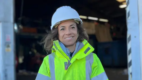 Faye Grime is wearing a white hard hat, and a High-vis jacket.
She is short and has medium length brown hair.