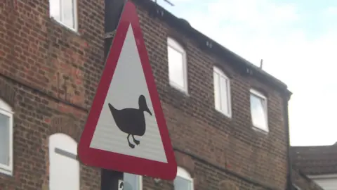 A triangular red and white sign attached to a pole. The sign features the silhouette of a duck.