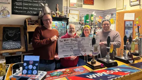 Mark Rogers from Peterborough Lions - wearing a brown jumper and blue jeans - next to Dave Williams wearing a red hoodie - standing next Carole Rogers from the charity - and Josh Barnard, staff member - all holding a big Wonky Donkey check 