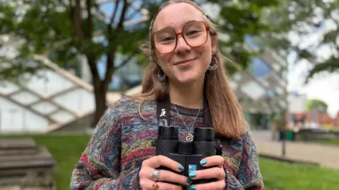BBC/Simon Thake A young woman with hair tied back and wearing a colourful jumper, holds a pair of binoculars