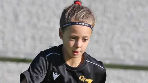 Family handout Poppy plays footballer. She has dark hair pulled back into a ponytail and is wearing a black football kit. She is looking down at the ground