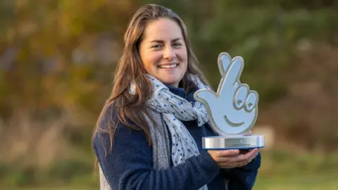 Phil Wilkinson Dr Cat Barlow smiling as she holds up a trophy in front of a blurred countryside background