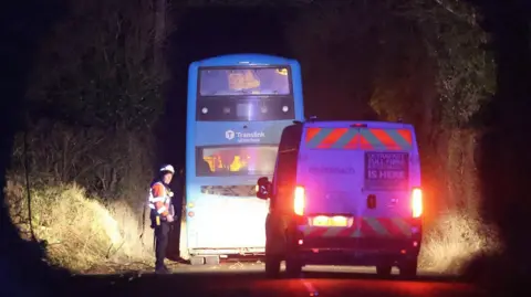 An emergency worker stands in front of a van and a Translink bus on a darkened road illuminated by the lights of the vehicles. There are trees on the edges of the road 