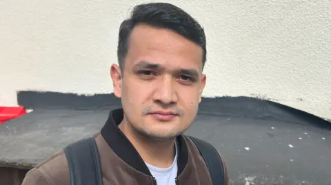 A man with dark brown hair combed in a side parting, wearing a brown velour bomber jacket over a white t-shirt, standing outside with a cream rendered building wall behind him.