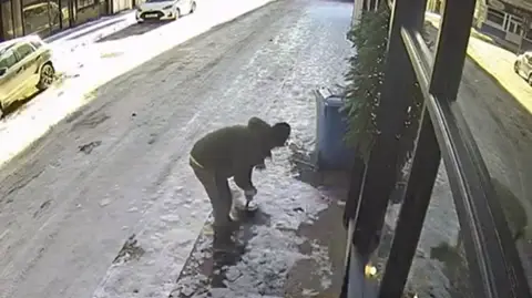 A screengrab of a CCTV recording showing a person wearing a big coat and a hat scooping up snow and ice outside a shop.