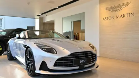 A silver Aston Martin car in a showroom. The sports car has a black number plate reading DB12 on the front.