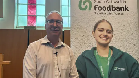 A man in a white shirt and black glasses, stood in front of a banner which says 'South Cotswolds Foodbank' and 'Together with Trussell'. There is a picture of a woman on the banner with a lanyard and fleece which says 'foodbank' on. The man is stood inside a church.