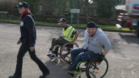 Shaun Whitmore/BBC A woman in black clothing and a baseball cap walking across a road, with two men in wheelchairs following behind her, one in hi-vis and dark clothing, the other in a grey hoodie and dark trousers. All three are wearing baseball caps. The blurred image of the back of a moving lorry can be seen to the right, and a road sign for Norwich and Fakenham. 