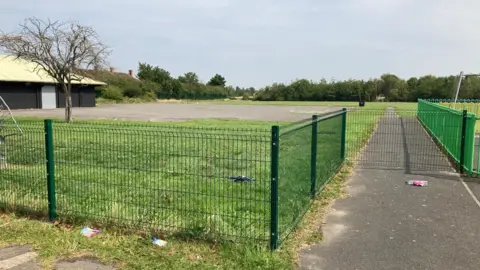 A green wire fence surrounds a field which is a mix of grass and concrete. There is a single-storey hut on the left and trees in the distance.