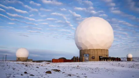 Getty Images Lanskap Arktik terpencil di Greenland utara, menampilkan Pangkalan Luar Angkasa Pituffik (sebelumnya Pangkalan Udara Thule). Gambar tersebut menunjukkan tiga kubah radar geodesik putih besar yang diposisikan di dataran tertutup salju. Kubah terbesar terletak di tengah struktur beton, dikelilingi kubah lainnya.