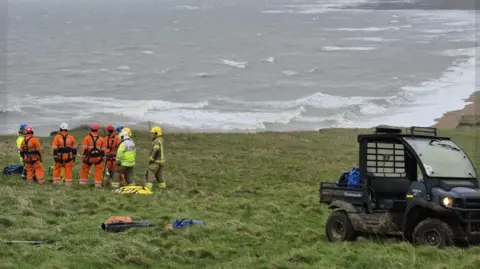 Bridport Fire Station Coastguard rescue teams in orange and yellow overalls standing at the top of a cliff working together to put in a plan to rescue a cockapoo who had fallen over the cliff.