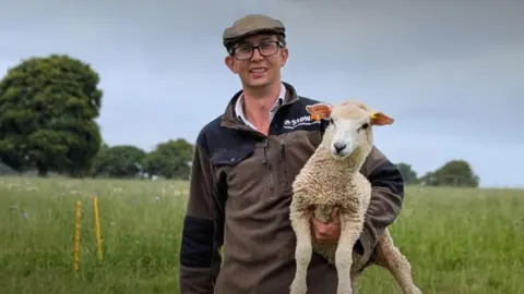 Farmer Karl Franklin stands in a field smiles for the camera whilst holding a lamb in the crook of his left arm. He's wearing a cloth farmer's cap, a brown and white outdoor jacket and black-rimmed glasses. 