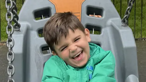 BBC A boy in a green jack and brown hair sits on a grey and orange ergonomic chair swing at a playpark.