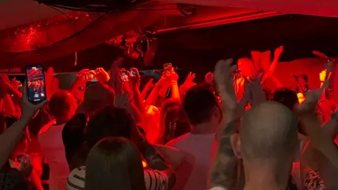 A crowd of people at a live music performance, holding up phones and hands under red lighting.
