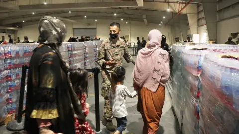 A soldier guides Afghan Special Immigrants. Pictured here are some women and children seen from behind, into the dining facility, August 20, 2021 at Camp As Sayliyah, Qatar
