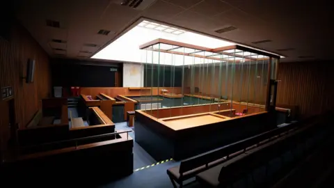 Bob Griffiths A square court room with a tall glass-panelled dock in the middle. The seating areas and wall cladding are wooden. A large skylight takes up most of the ceiling.