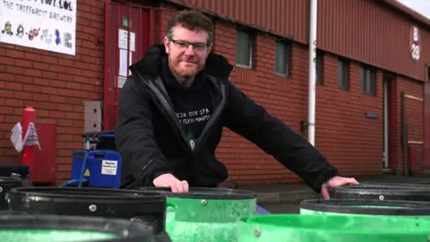 Philip Thomas, wearing glasses and a dark jacket, kneels behind a row of green beer barrels outside his brewery in Treforest. He is smiling at the camera, while the brick building housing his brewery is immediately behind him.