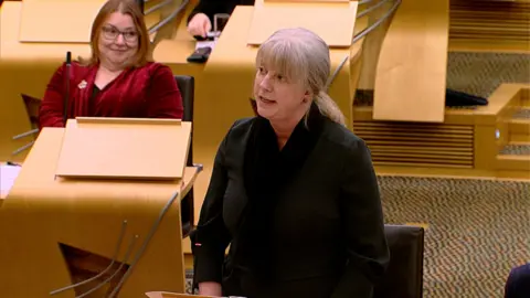 Shona Robison stands to deliver the Scottish Budget in the Scottish Parliament, her light-coloured hair is tied in a pony tail and she is wearing a black dress