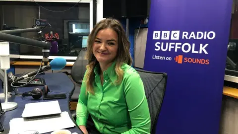 Vikki Irwin/BBC A woman is sitting in the radio studio at BBC Radio Suffolk. She has long hair and is wearing a bright green shirt. 