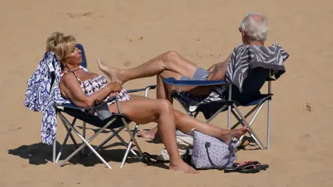 PA Media Two people sit on deckchairs on a sandy beach. They've got their eyes shut and feet up, wearing bikini's and swim shorts.