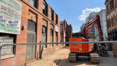 A digger in the middle of the street working near a factory.