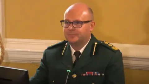 Dawn Sturgess Inquiry Mr Darch wearing a green military uniform with coloured patches. He is wearing dark framed glasses, has a bald head and is clean shaven. He is looking to the left of the camera at Mr Mansfield. 