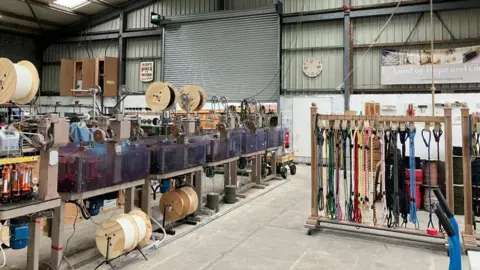 The inside of a ropemaking warehouse, containing ropemaking machinery resembling wooden bobbins. An array of dog leads are hanging from wooden stands.
