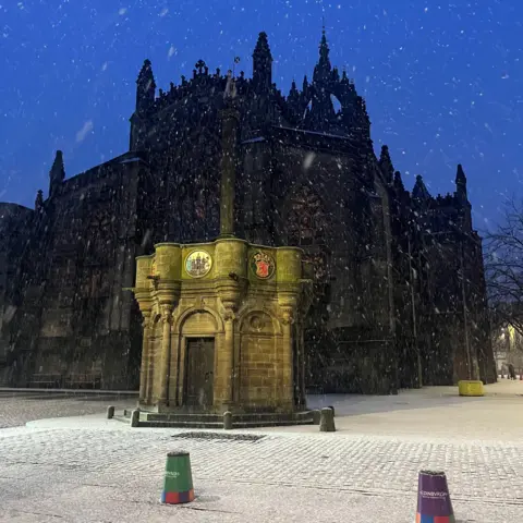 Helen Mcvey Snow falls over a historic monument in front of a Gothic-style building, as evening light casts a deep blue hue across the sky. 