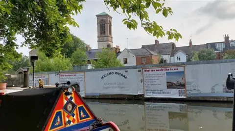 A view of the derelict site with tattered banners alongside the canal in Jericho. The church can be seen in the background. There is also a boat in the water.