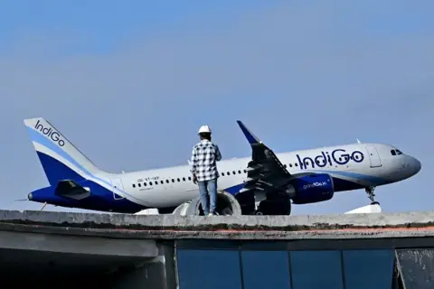 AFP via Getty Images A man stands atop an under-construction building as an Indigo aircraft prepares to land at Kempegowda International Airport in Bengaluru on December 4, 2025. 