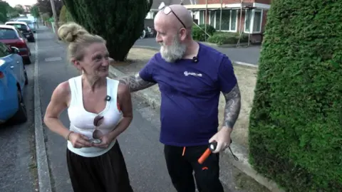 A woman smiling at a man who has his arm around her. They are stood on a residential street.