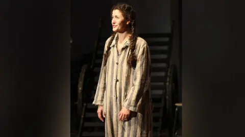 Voices of the Holocaust The actor playing Susan stands speaking to the audience, wearing a striped nightshirt.