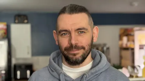 Stephen Browne with short hair and a beard and wearing a blue hoodie and white t-shirt smiles at the camera. In the background you can see kitchen cupboards and utensils.