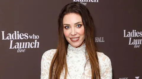 Getty Images A woman in her 40s is wearing a pink lipstick while lace dress as she poses in front of a 'ladies who lunch' sign. 
