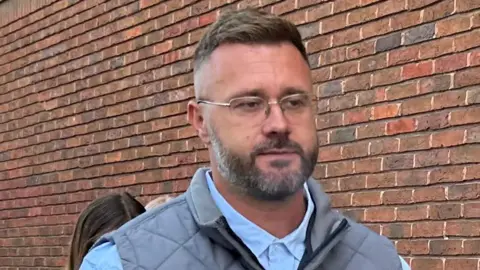 PA Media A man with short, light brown hair and brown/grey facial hair. He has glasses and is wearing a blue shirt and grey gilet. He is walking in front of a brown brick wall