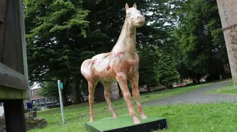 North Lincolnshire Council A brightly painted, life-sized sculpture of a horse stands on a patch of grass. Red handprints and birds are among the designs on the horse. 