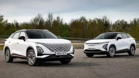 Chery Two white Chery Omoda cars sitting at right angles to each other on a tarmac. Beyond them is a barrier and beyond that are trees.