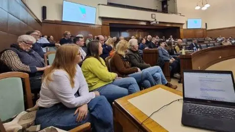 Aled Thomas A group of people sit in the public gallery in a council chamber. They are watching members of the committee talk, and plans for the site can be seen behind them faintly on screen. 