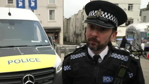 A police officer stood next to a police van. They are both stood in a small city centre plaza.