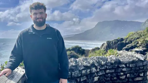 Lloyd Lloyd, who has dark curly hair and facial hair, is wearing a dark jumper and is standing by a stone wall at a coastal viewpoint, with ocean waves, cliffs, and a cloudy blue sky in the background.