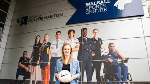University of Wolverhampton Kerys Harop is standing holding a football in front of a large sports‑themed mural outside the Walsall Sports Centre at the University of Wolverhampton. She has blonde hair and is wearing a denim jacket over a black top
