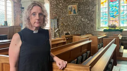 Tom Larsen-Wright/BBC Jo Parrott wears a black top and a white dog collar, as she leans against a pew in a medieval looking church, with stone walls and stained-glass windows.