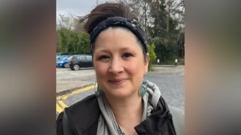 Daniela Slater has dark hair tied up and is wearing a headband. She is wearing a grey hooded top and black jacket, blue earrings and nose ring and lip piercing.