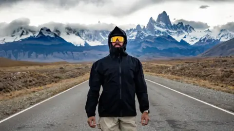 Charlie Probets Mike Humphreys stood in the middle of the road looking at a camera. He is wearing beige shorts, a black coat with its hood up and yellow running glasses. Behind him are mountains with clouds looming over. 