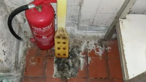 Gloucester City Council A red fire extinguisher next to a yellow piece of plastic on a dirty tile floor. It is unclear what is on the floor but it is black, grey, brown and white in colour. The walls, which should be white, have brown and black marks all over them.