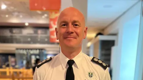 A man in a fire service uniform in an office. He is smiling and look straight at the camera.