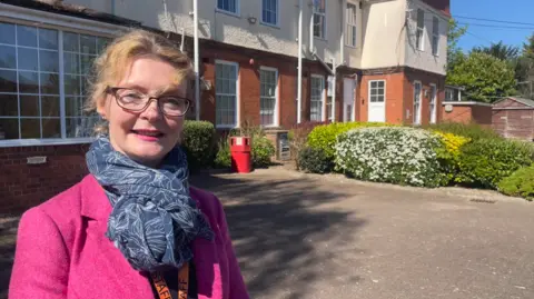 A woman in a pink coat and blue and white scarf is wearing glasses and standing on the left side of the photo. A building, which has some bushes in front of it, is behind her.